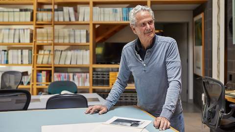 Don Chadwick, in camicia blu, accanto a un tavolo nel suo studio. La parete alle sue spalle presenta scaffali pieni di libri e raccoglitori.