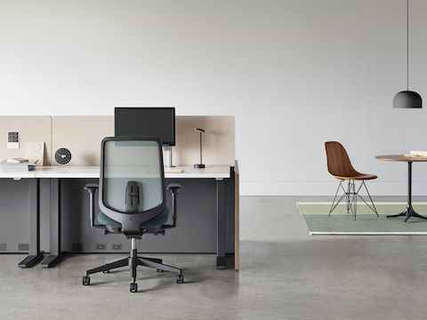 A black Verus chair with a black Motia Sit-to-Stand Table and Canvas Channel workstation with a Nelson Pedestal Table and Eames Moulded Wood Side Chair in the background.
