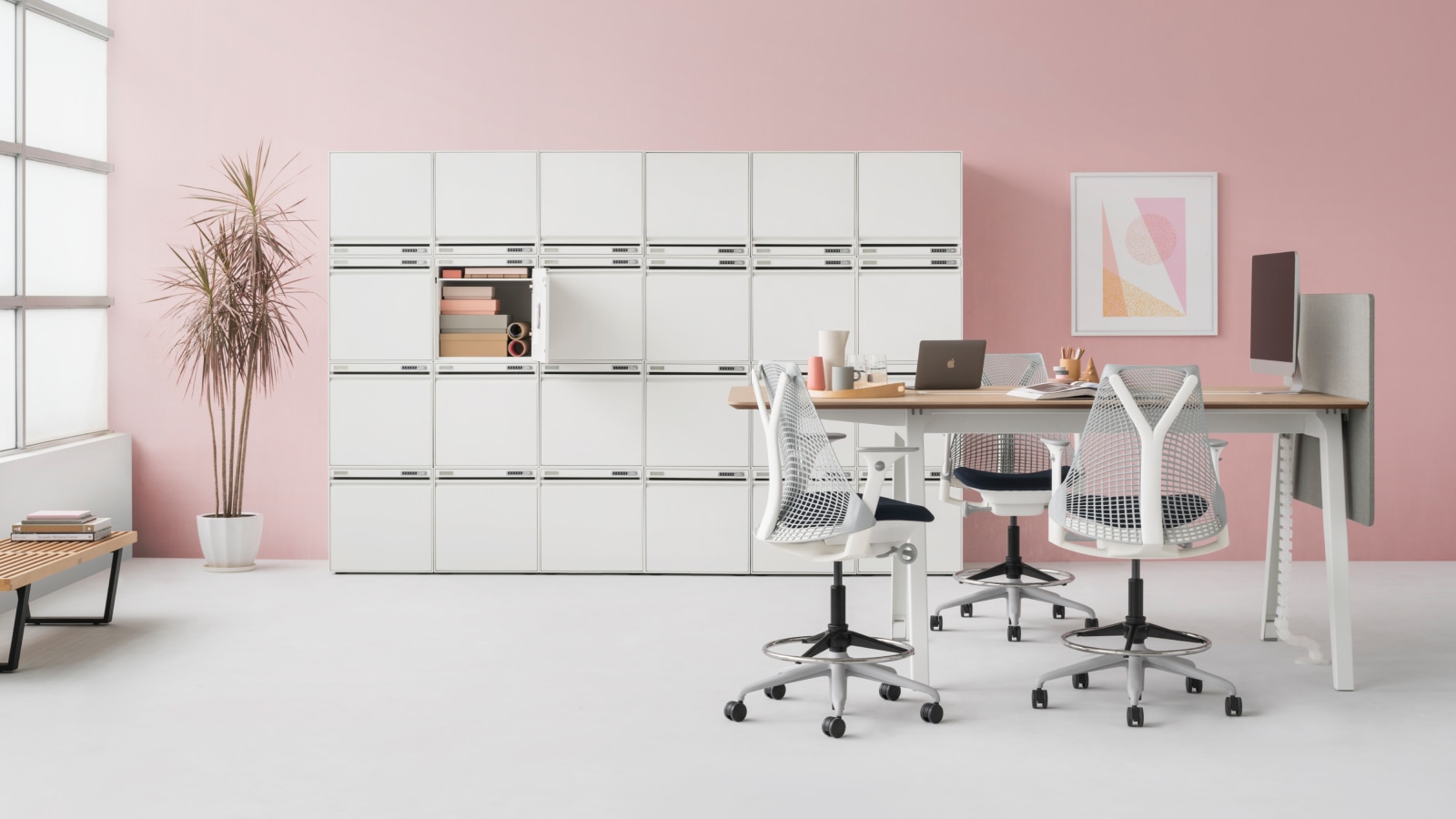 A six-piece CKL Keyless Storage Unit sits against a pink wall. It features 24 small storage lockers. In the foreground, a high Optimis Table and four Setu stools combine to form a meeting area.