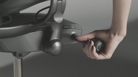A black Aeron Chair with a person sitting in it, reaching down to adjust the tilt tension control knob.