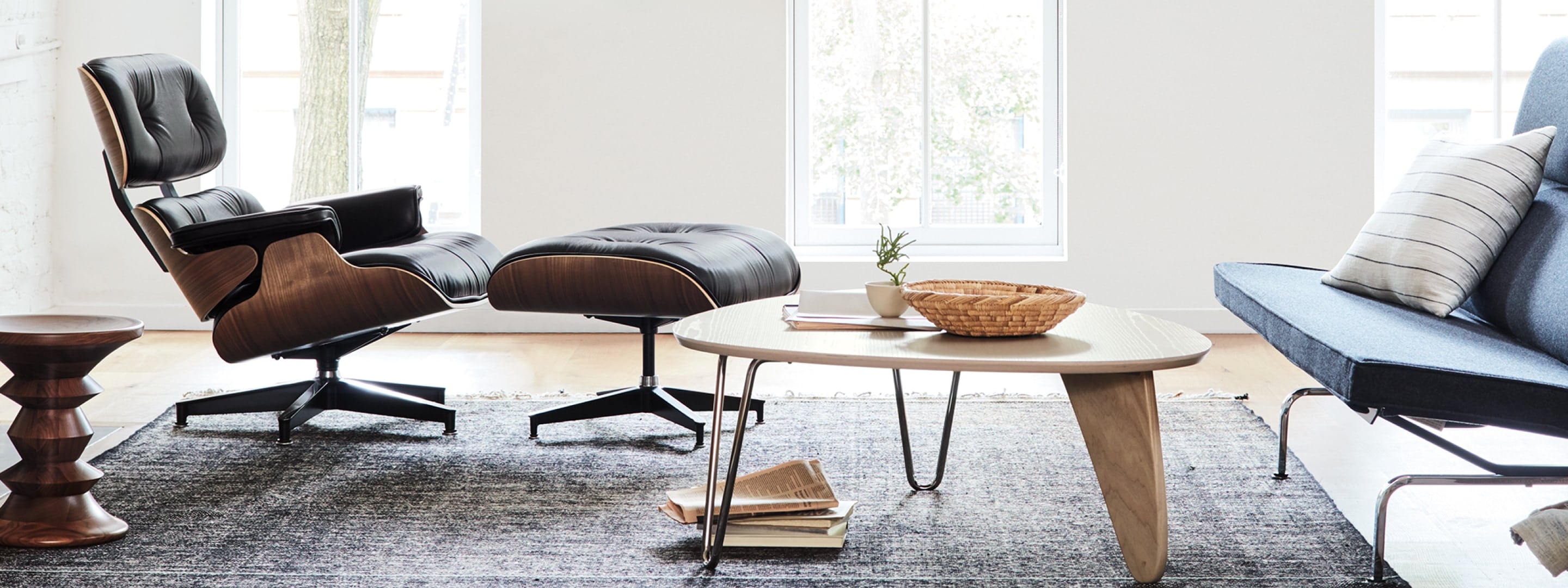 An Eames Lounge Chair and Ottoman, Noguchi Rudder Table, Eames Sofa Compact, and Eames Walnut Stool in a living room setting with large windows in the background.